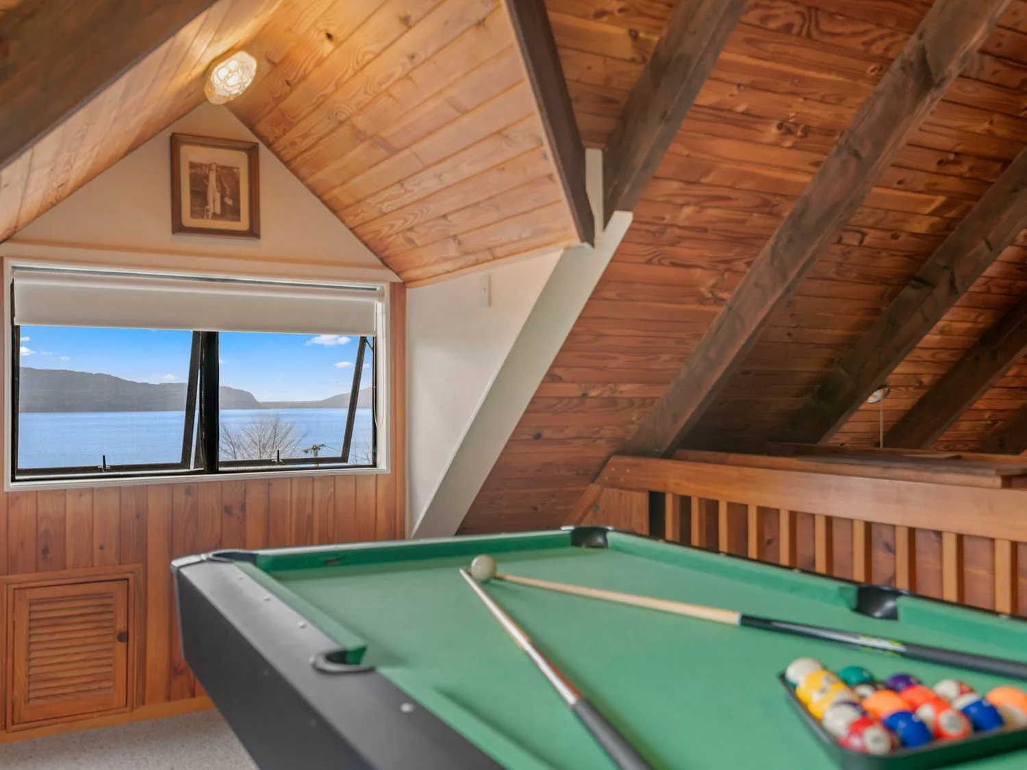 A pool table with cue sticks in a wooden loft room with a window showing a lake view at Splendour on Spencer - Lake Tarawera Holiday Home