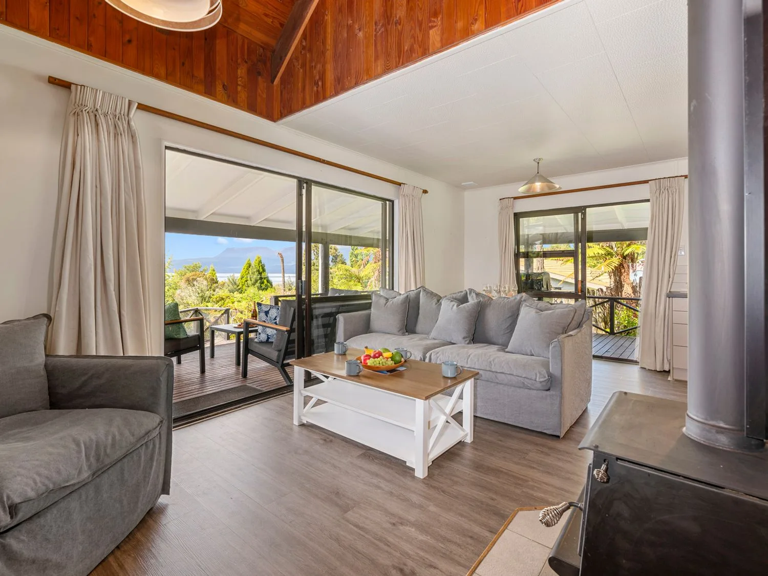 A living room with gray sofas a wooden coffee table with fruit a wood stove and sliding glass doors leading to an outdoor seating area at Splendour on Spencer - Lake Tarawera Holiday Home