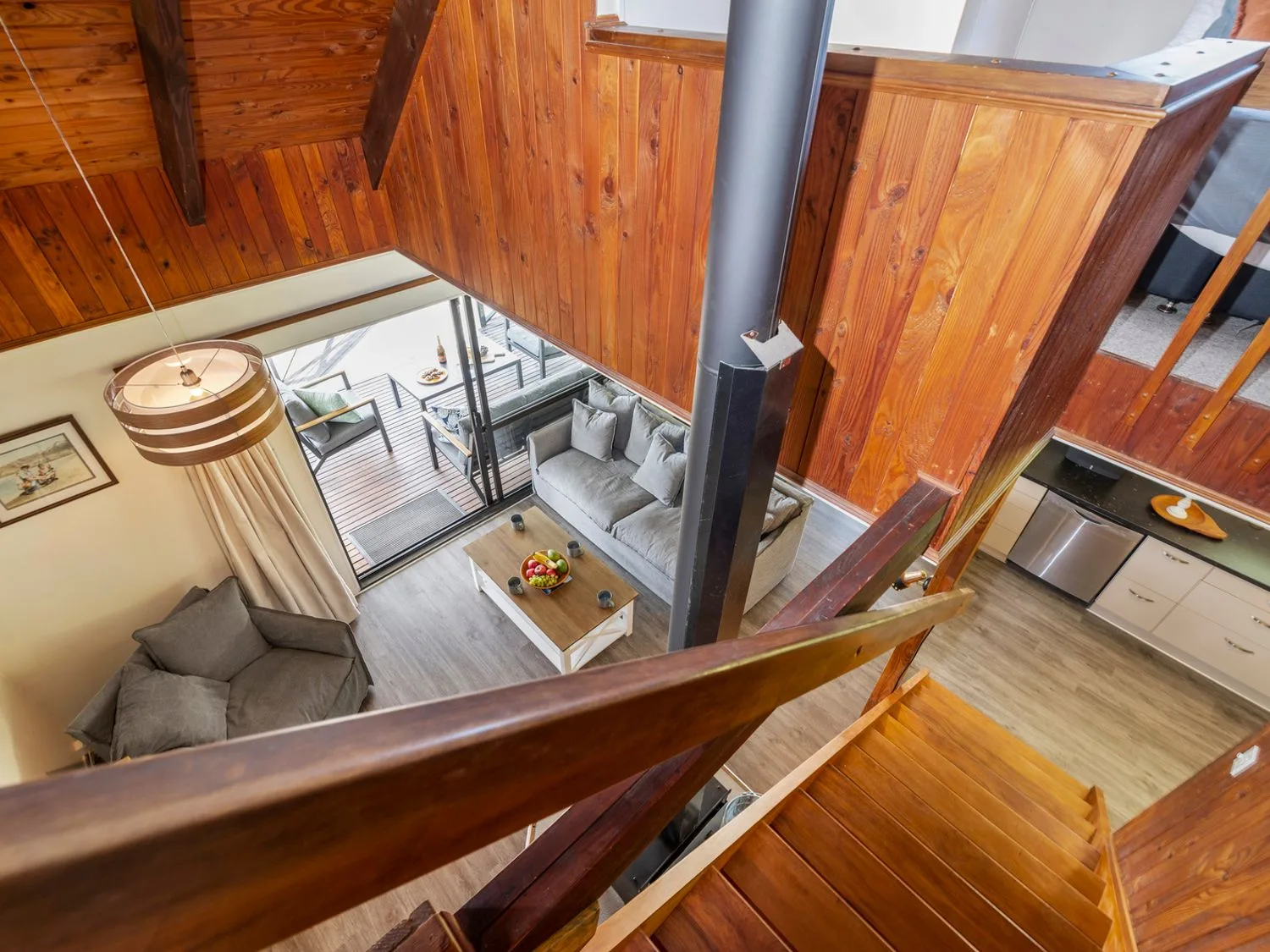 View down wooden stairs to a living room with sofas and a coffee table beside a glass door leading to an outdoor deck at Splendour on Spencer - Lake Tarawera Holiday Home
