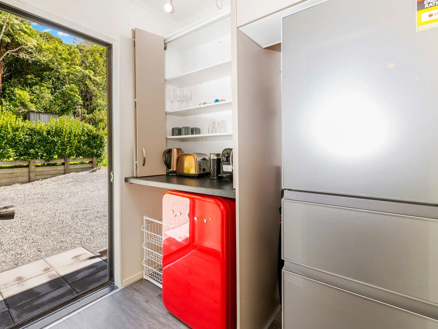 A kitchen area with an open cupboard containing a kettle toaster coffee machine and glasses a bright red mini fridge under the counter and a silver refrigerator next to a door leading outside with greenery visible