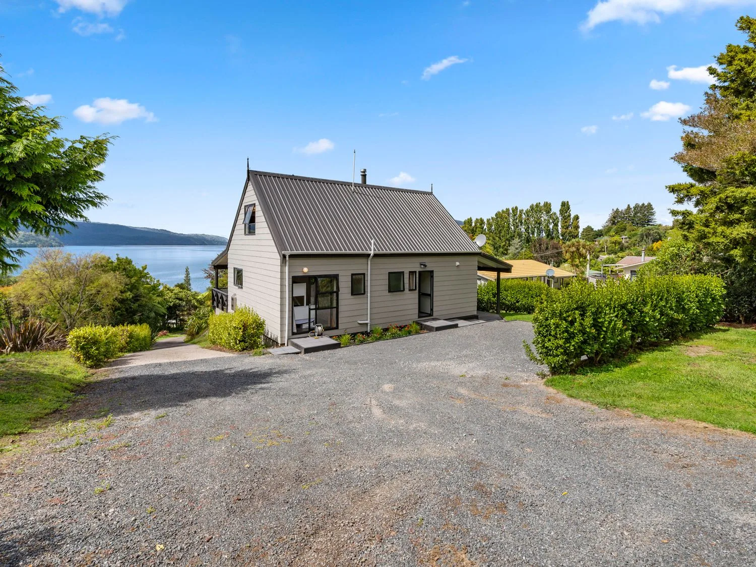 A house beside a gravel driveway, with a lake and trees in the background.