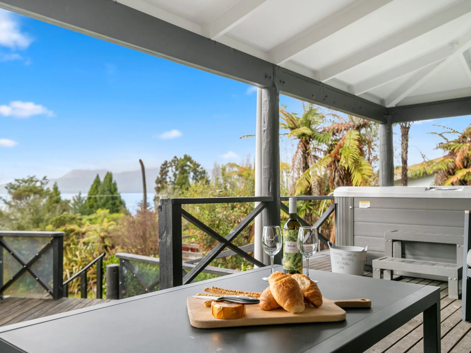 A covered outdoor patio with a serving board and wine glasses overlooking trees and water.