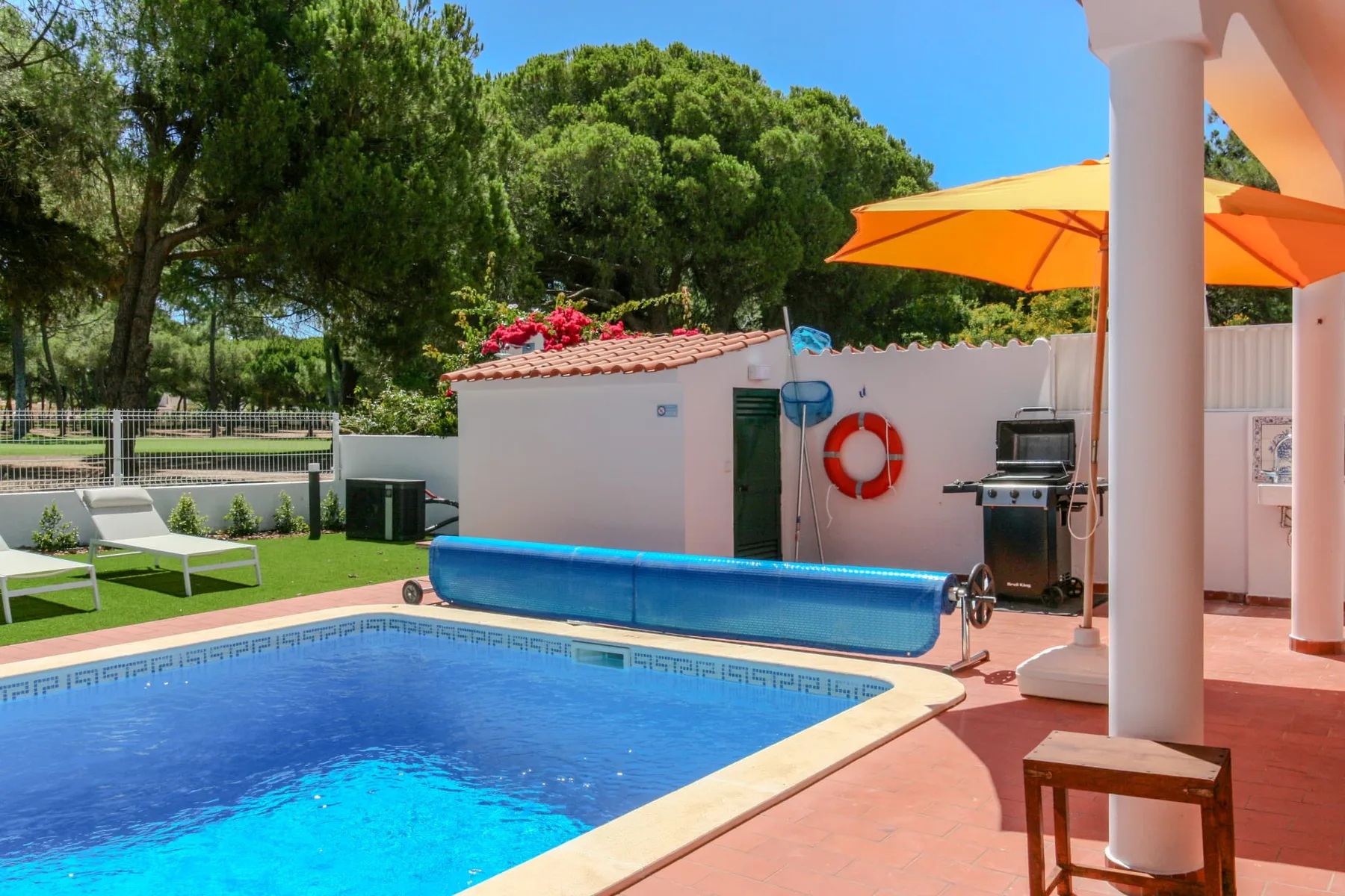 An outdoor swimming pool beside a patio with an orange umbrella and seating.