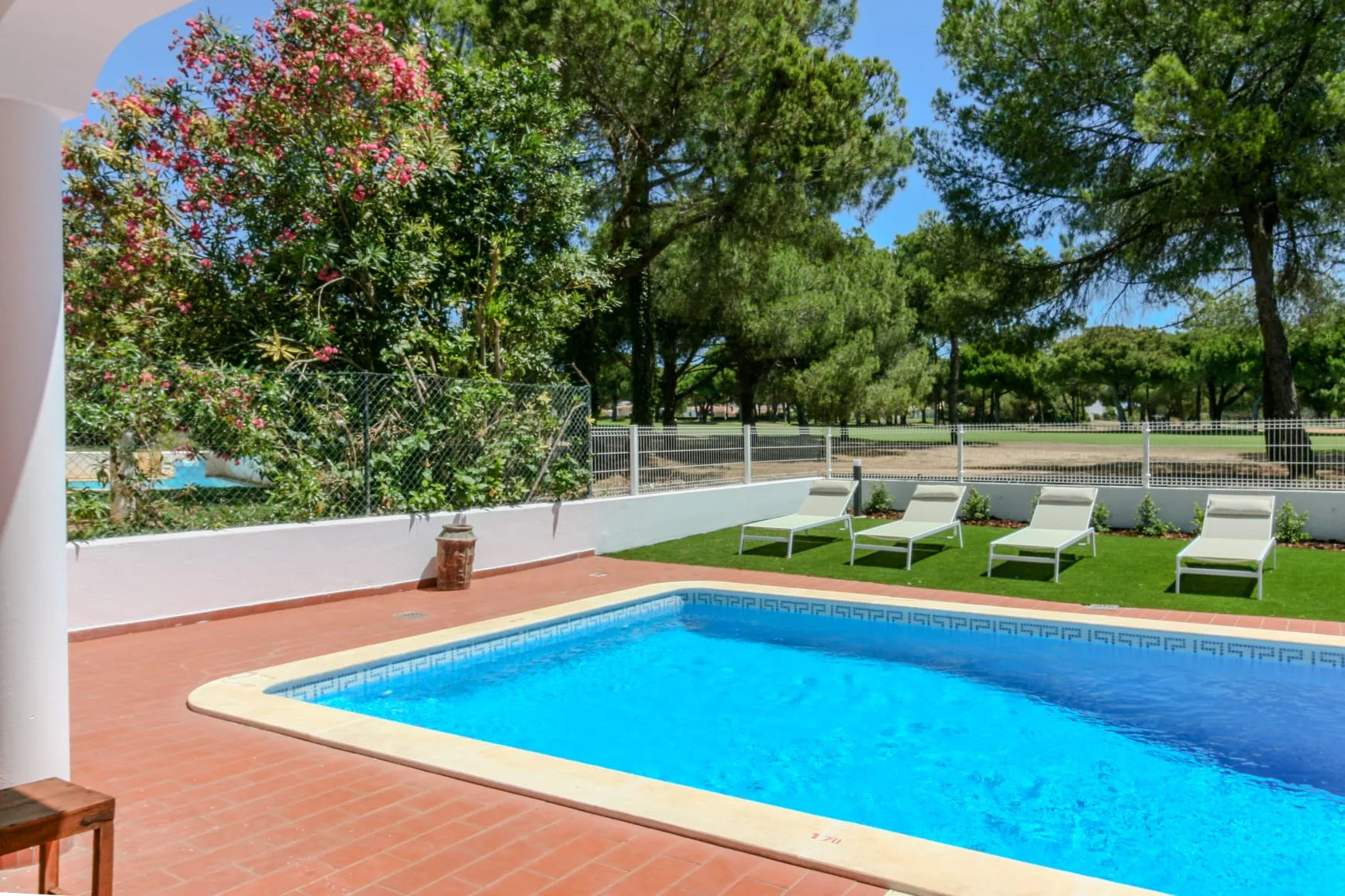 An outdoor swimming pool sits beside a tiled patio, with lounge chairs and trees beyond.
