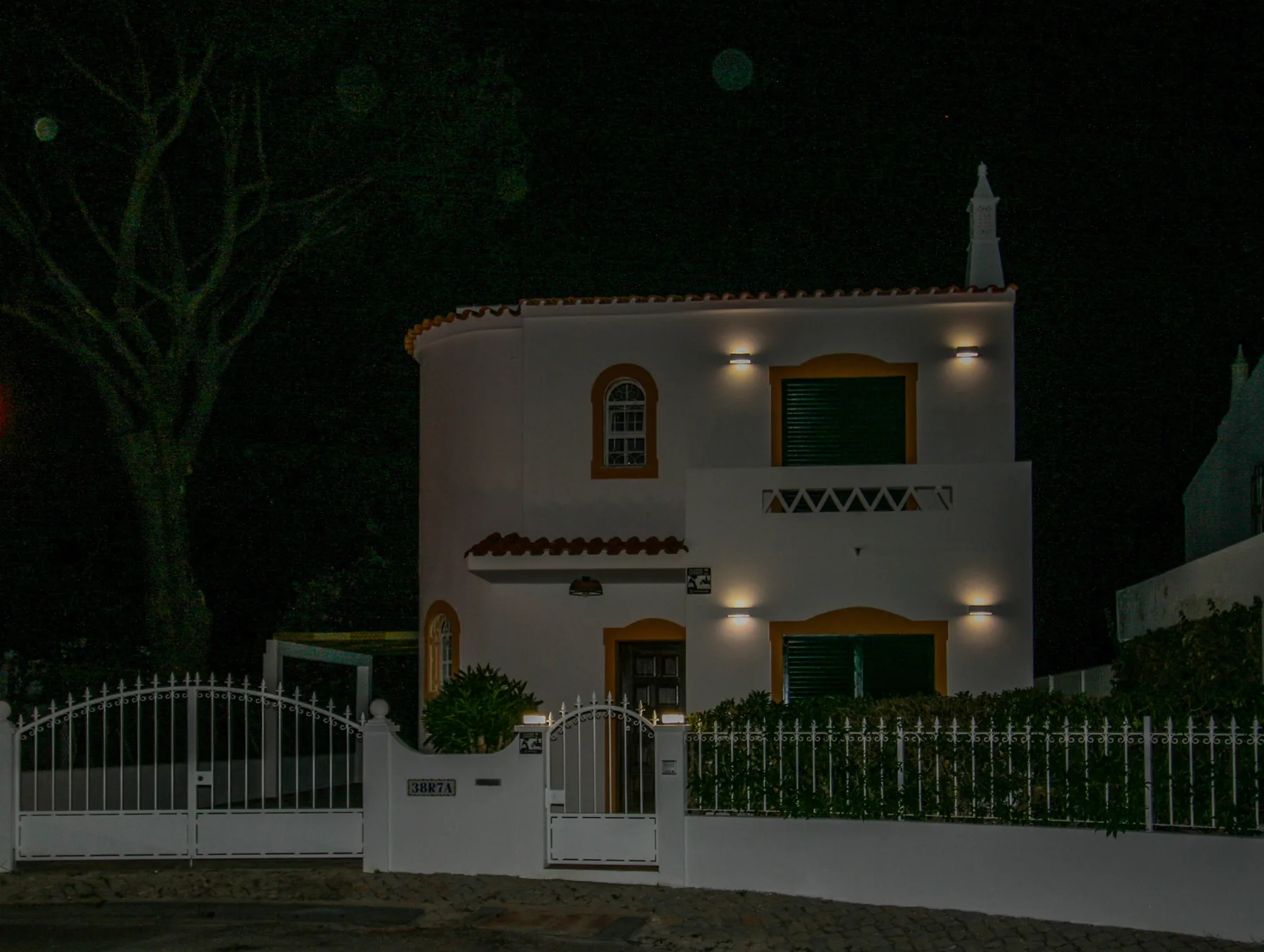 A two-story villa stands behind a white fence at night, with exterior lights on.