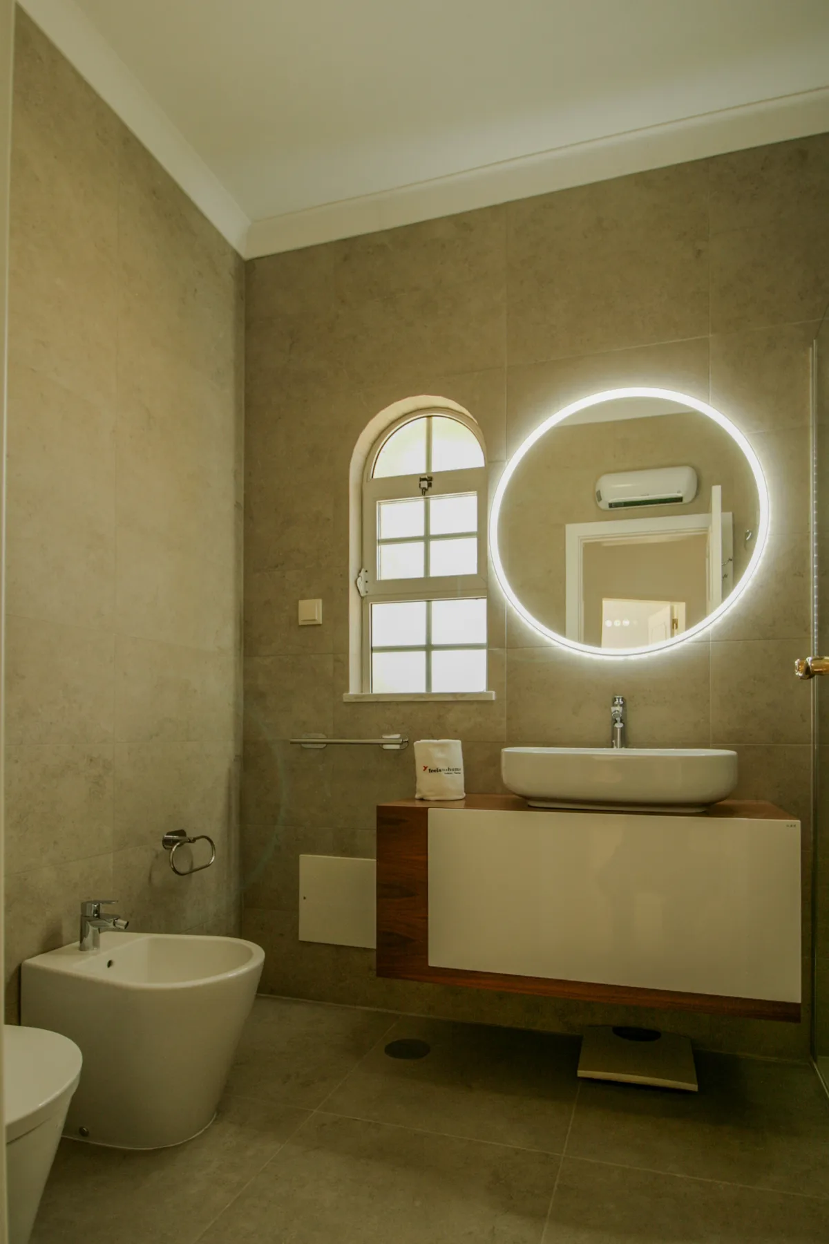 A tiled bathroom with a floating vanity, vessel sink, and illuminated round mirror.