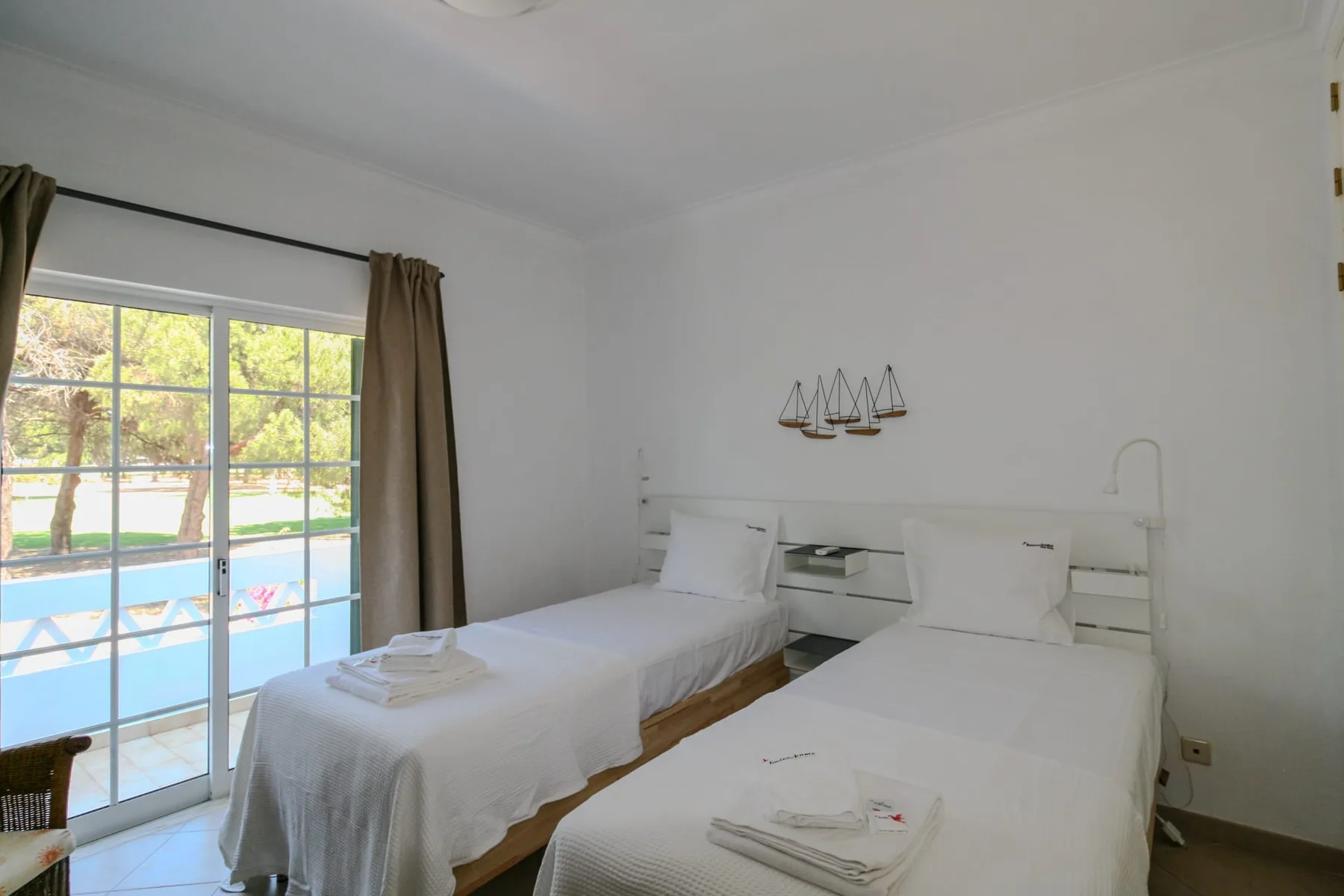 A bright bedroom with two beds beside a large window overlooking a pool.