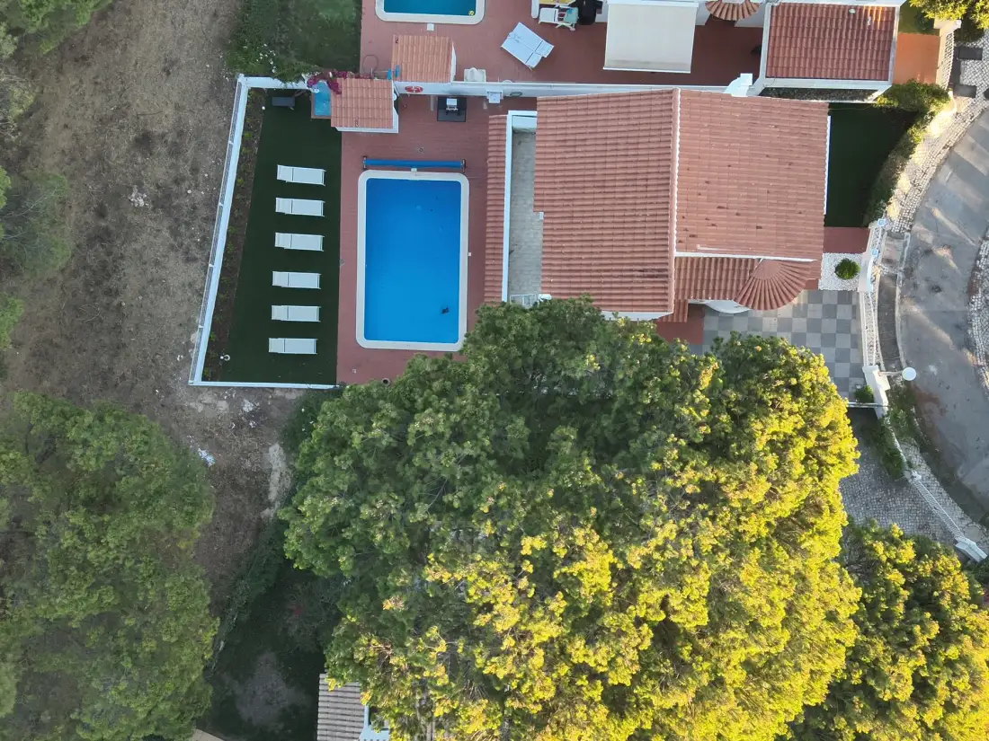 An aerial view shows a villa beside a rectangular swimming pool and surrounding trees.