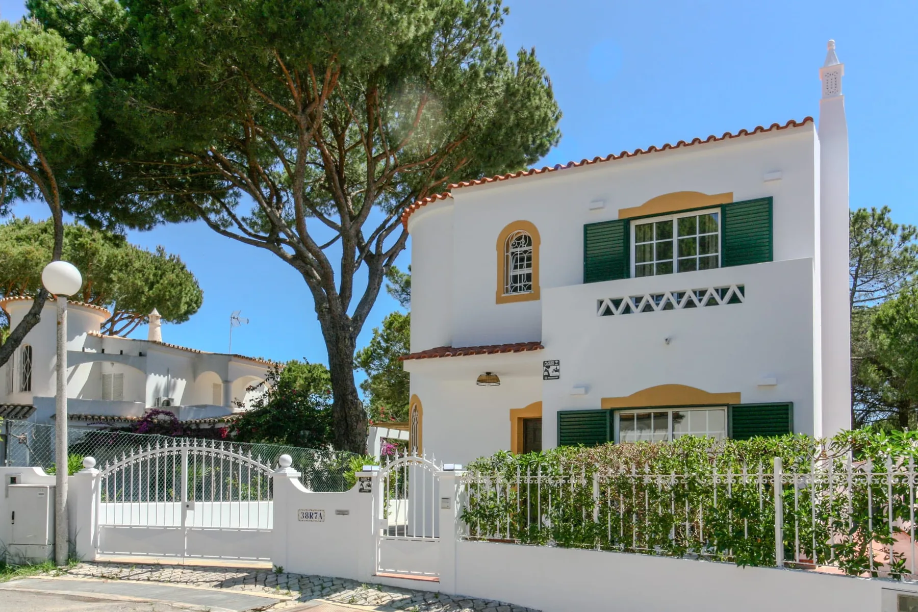 A white two-story house stands behind a gated entrance and leafy trees.