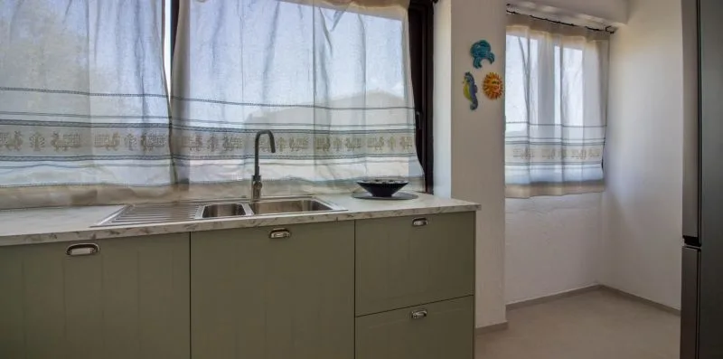 Una encimera de cocina con fregadero queda bajo ventanas con cortinas transparentes.