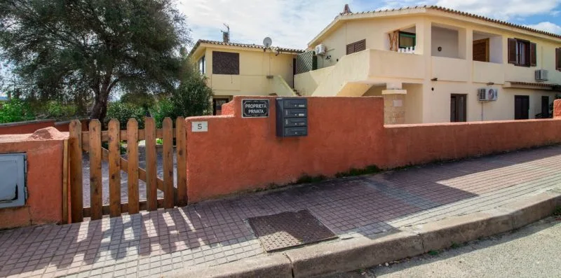 Un edificio claro de dos plantas se alza tras un muro rojizo y una puerta de madera.