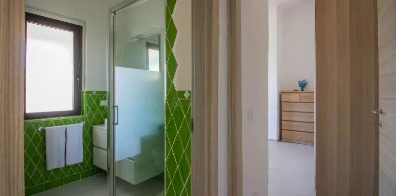 Un baño con paredes de azulejos verdes, una ducha de cristal y una toalla colgada bajo la ventana.