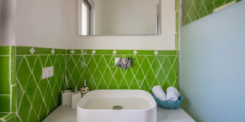 Un lavabo de baño está bajo un espejo, junto a azulejos verdes decorados.