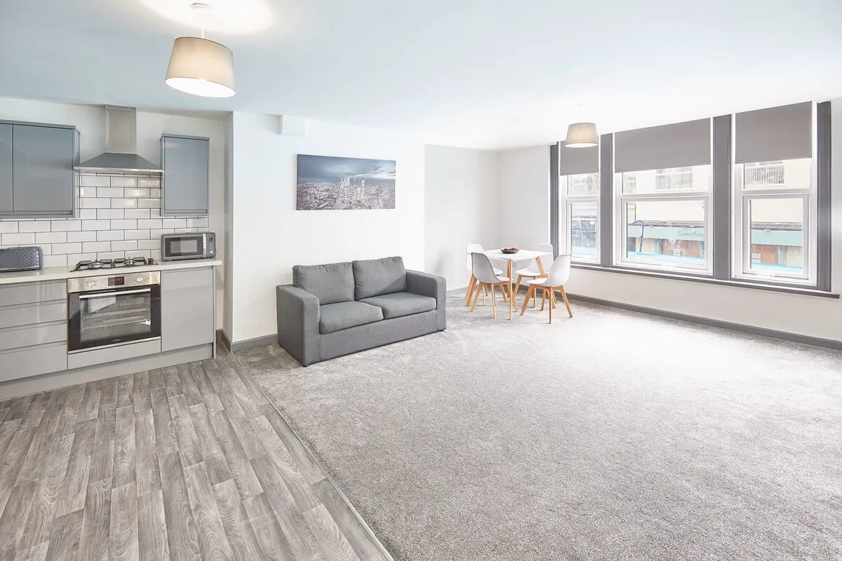 An open-plan living area with a compact kitchen, gray seating, and a dining table by the windows.