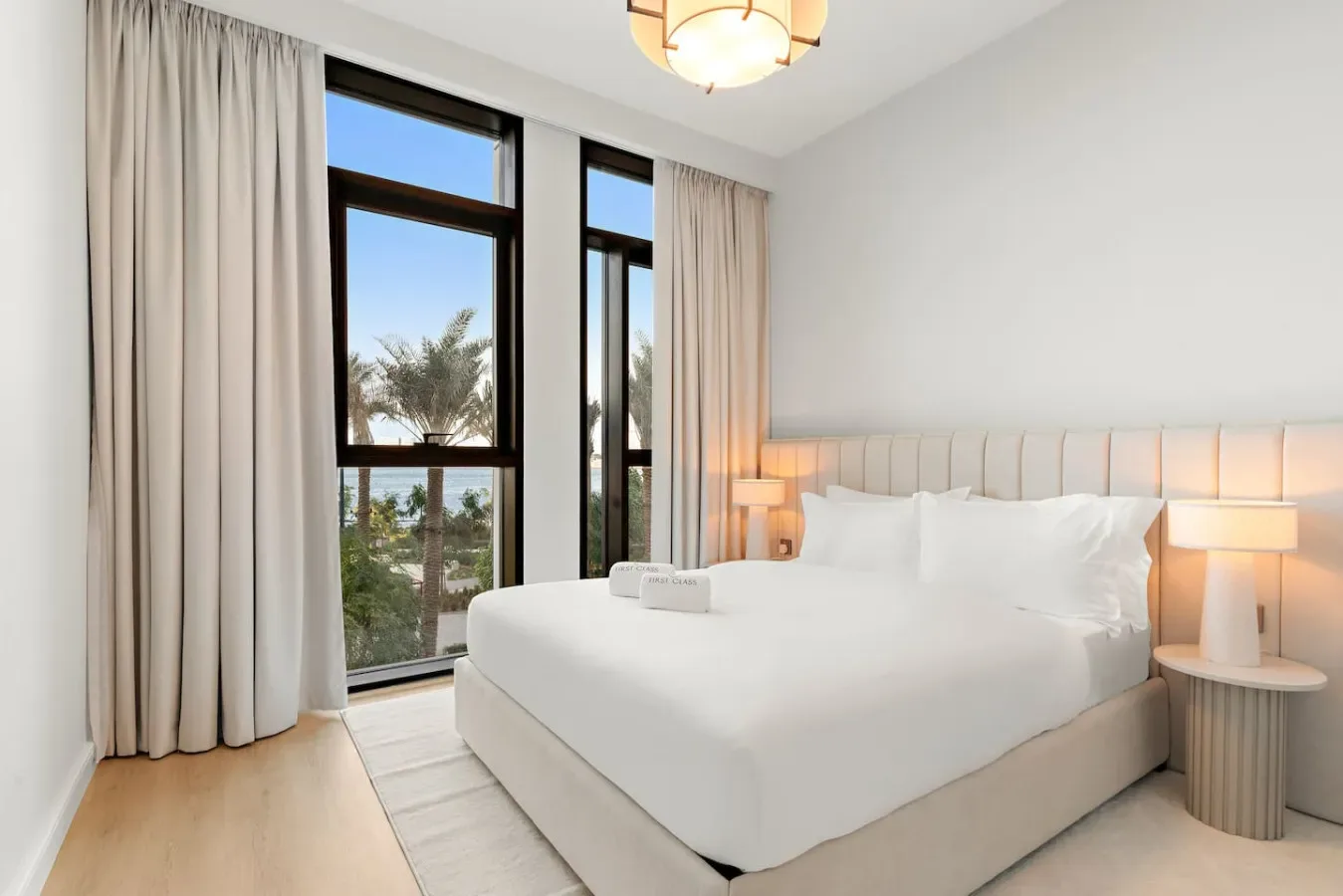 Un dormitorio luminoso con una cama bien tendida junto a ventanas altas con cortinas.