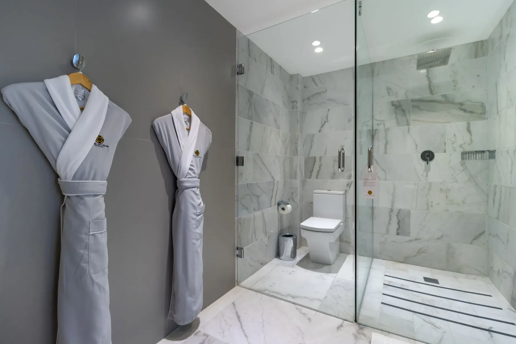 Baño con albornoces grises, baldosas de mármol y una ducha de cristal.