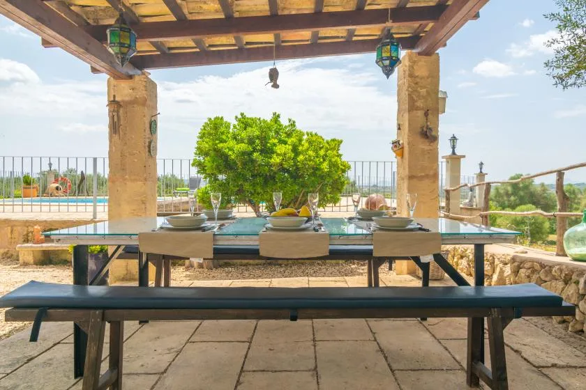 Una mesa de comedor al aire libre está bajo una pérgola de madera junto a una piscina.