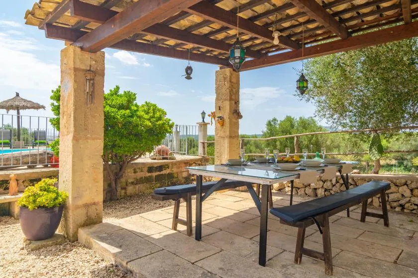 Una zona exterior de comedor a la sombra, con una mesa larga bajo una pérgola de madera.
