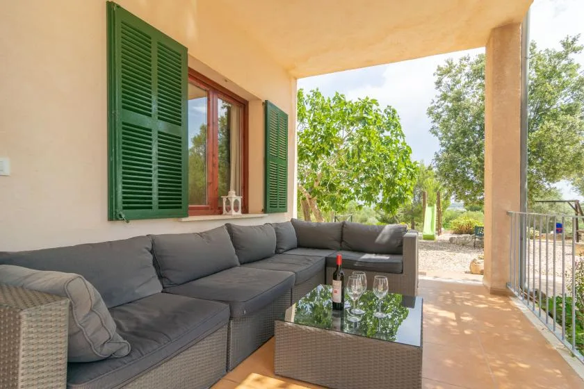 Un patio cubierto tiene asientos acolchados y una mesa con tapa de vidrio junto a un jardín abierto.