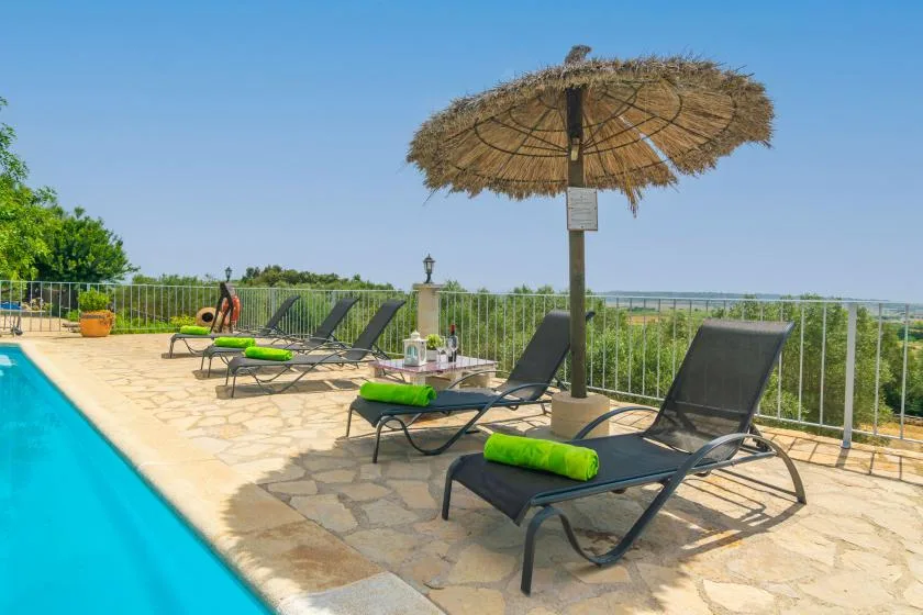 Una terraza soleada junto a la piscina, con asientos reclinables y una sombrilla de paja.
