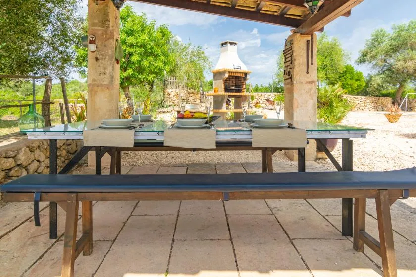 Comedor exterior cubierto con una mesa larga frente a una barbacoa de piedra.