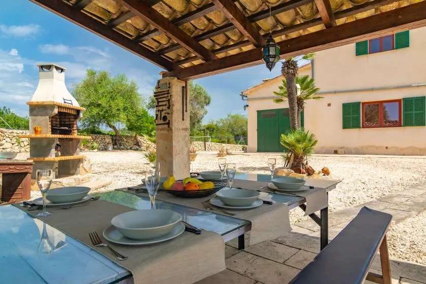 Una zona de comedor al aire libre está bajo una pérgola de madera, junto a una casa y una barbacoa de piedra.