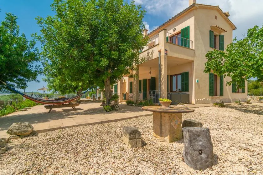 Una casa de dos plantas se alza junto a un patio de grava con una hamaca y árboles.