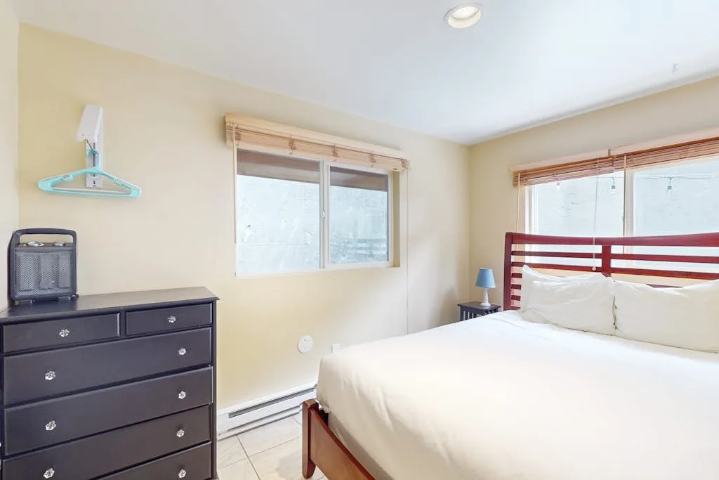 A bedroom with a bed, a dark dresser, and windows with blinds.