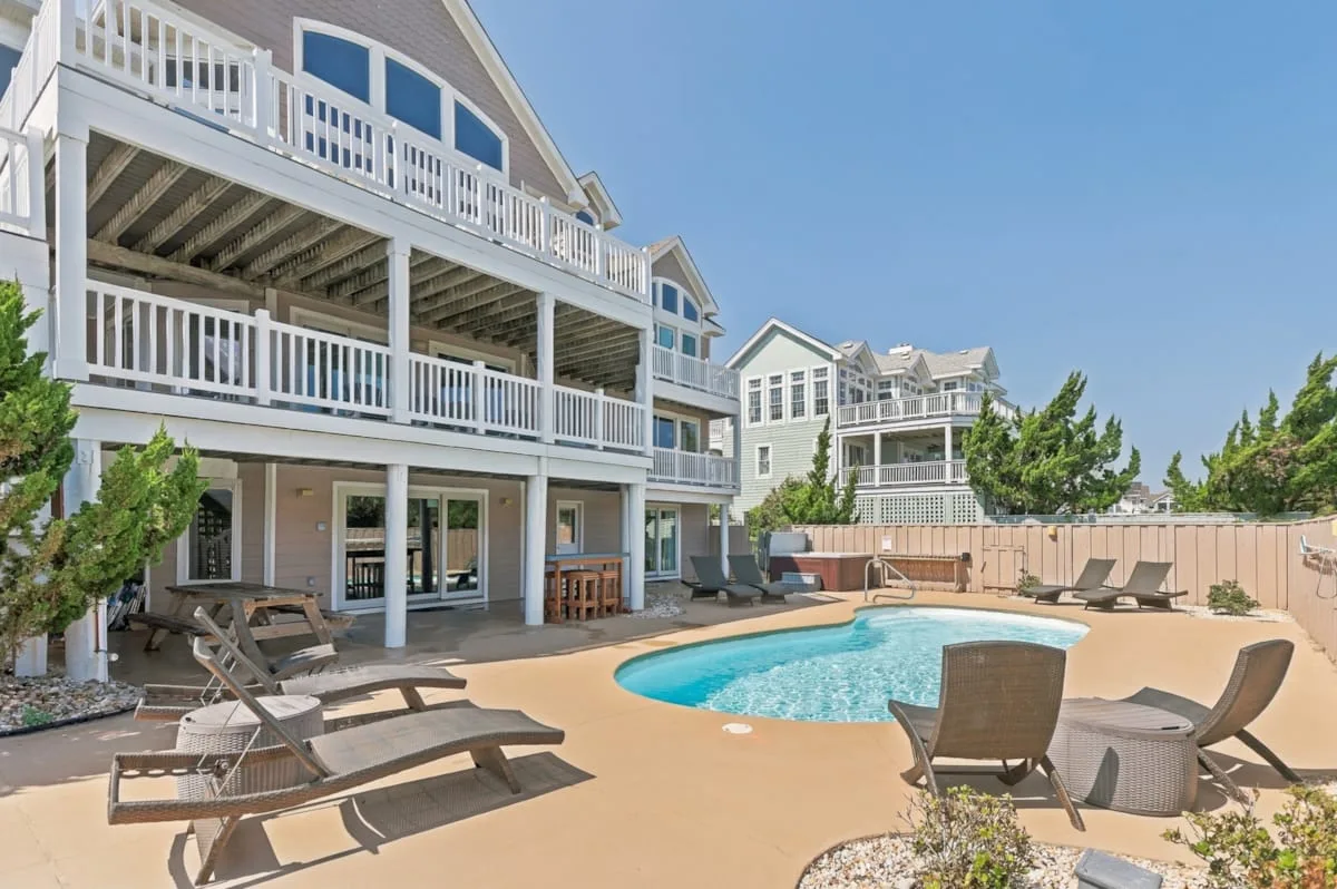 A backyard swimming pool sits beside a large house with balconies and patio seating.