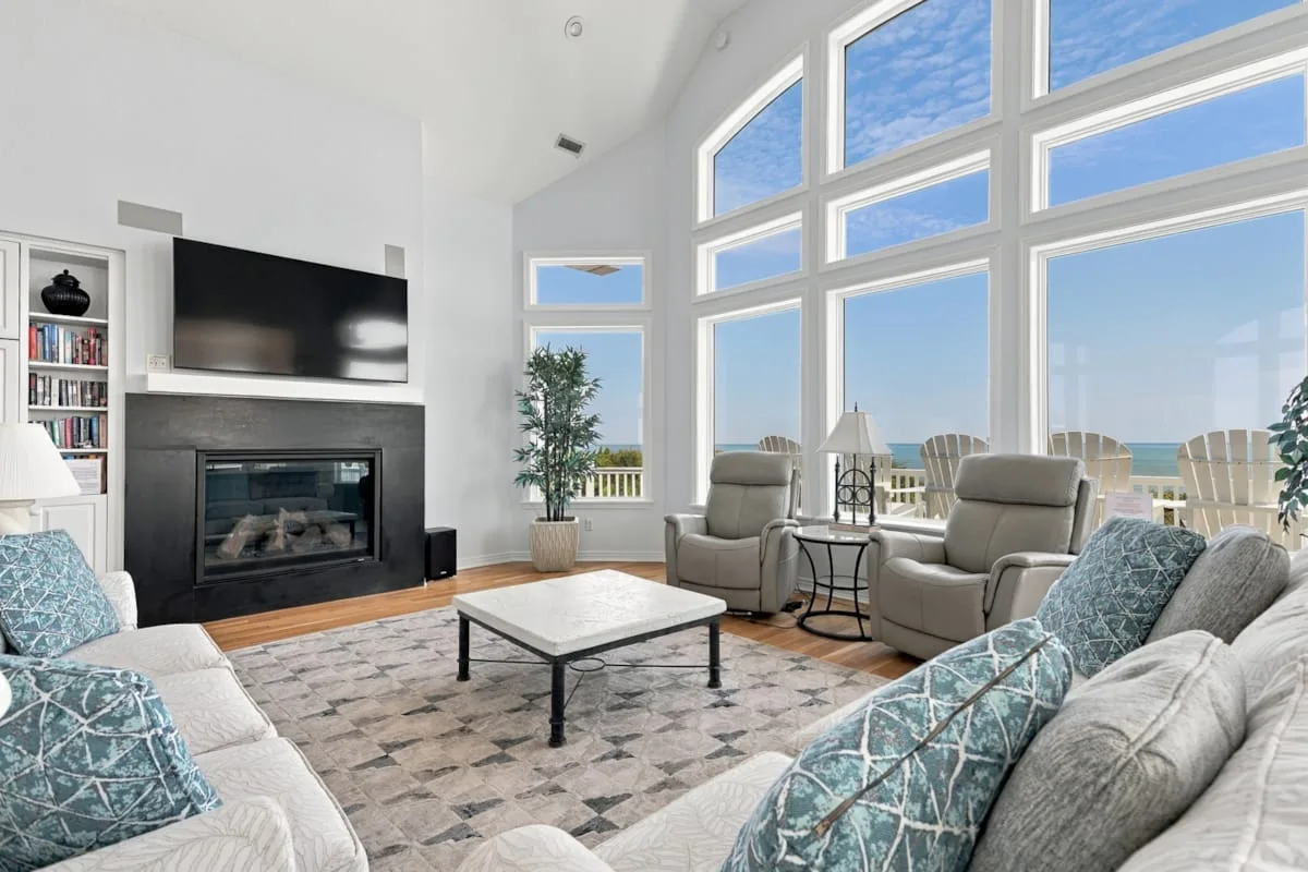 A bright living room with seating, a fireplace, and large windows overlooking the water.
