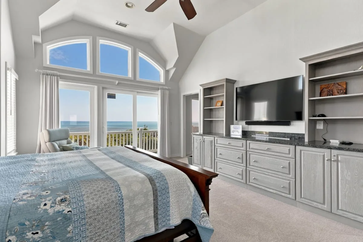 Un dormitorio luminoso tiene una cama grande junto a ventanas altas con vistas al océano.