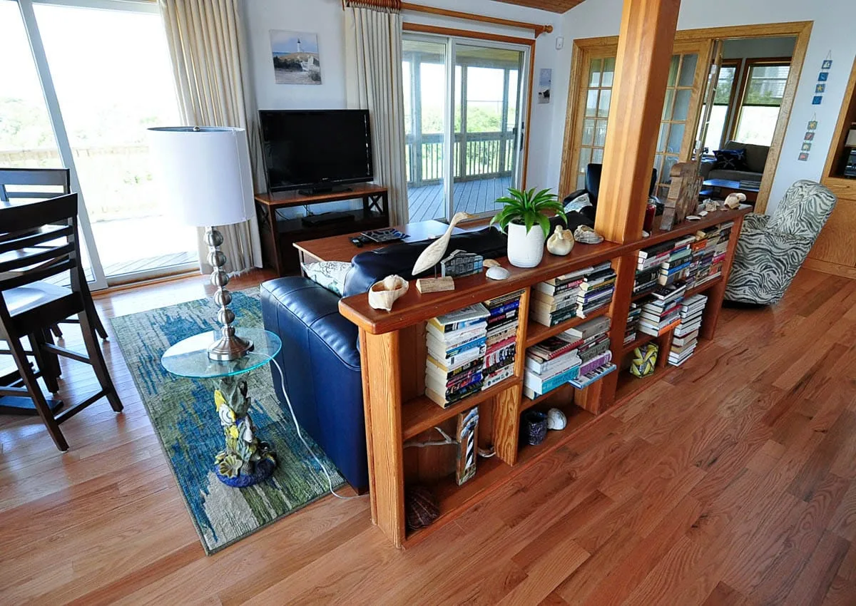 Una sala luminosa con televisor, asientos y un separador lleno de libros.