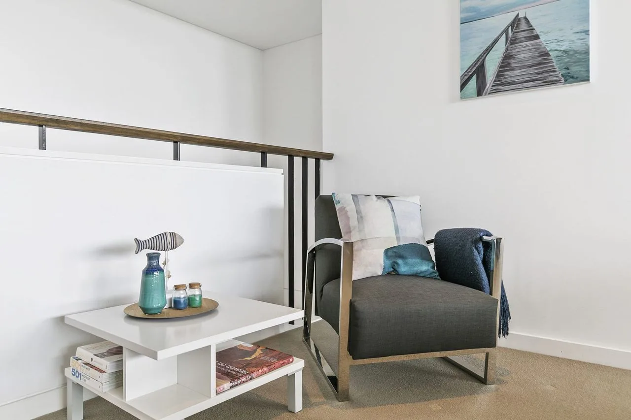 Una zona de estar luminosa con un sillón oscuro, cojines y una mesa blanca baja.