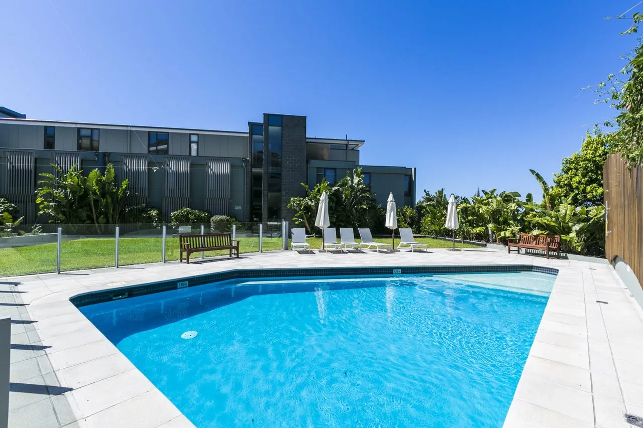 Piscina exterior junto a un edificio moderno y una zona ajardinada con asientos.