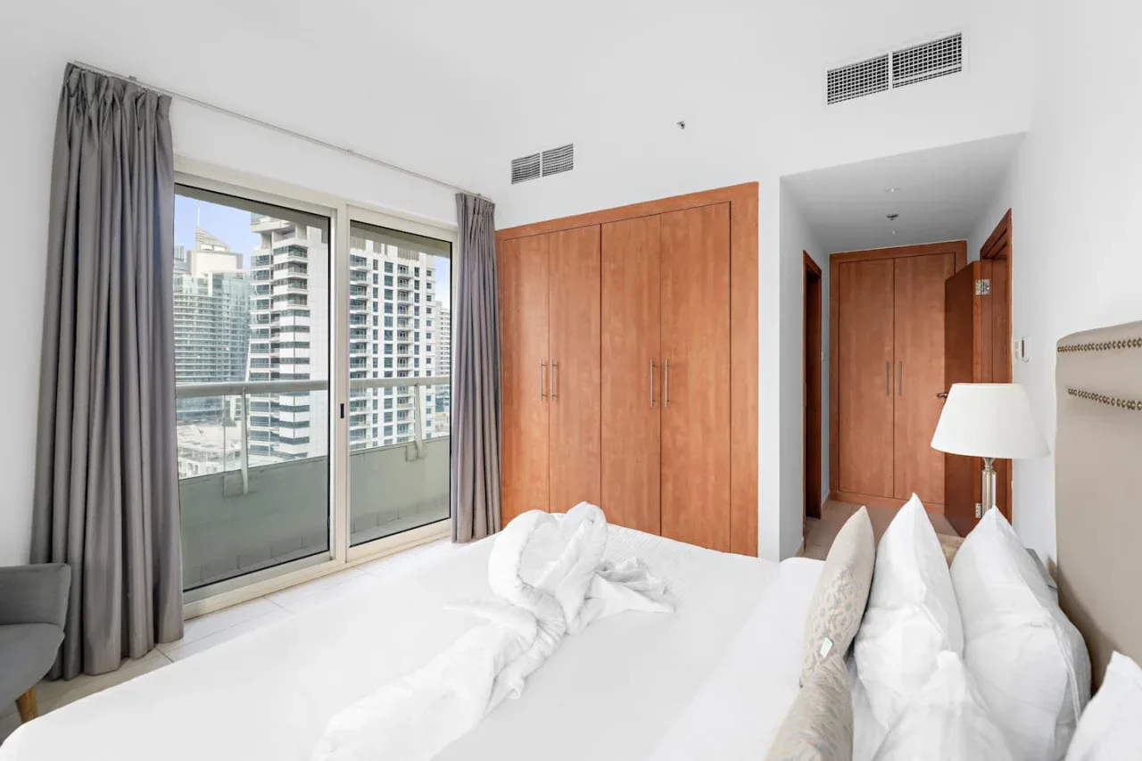 A bright bedroom with a large window overlooking nearby high-rise buildings.