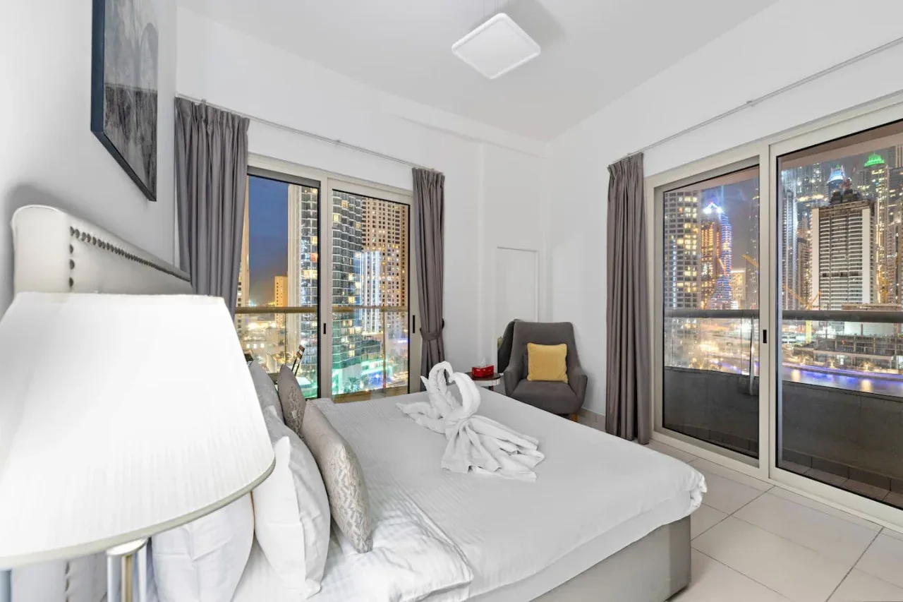 A bedroom with a neatly made bed, seating, and large windows overlooking city lights.