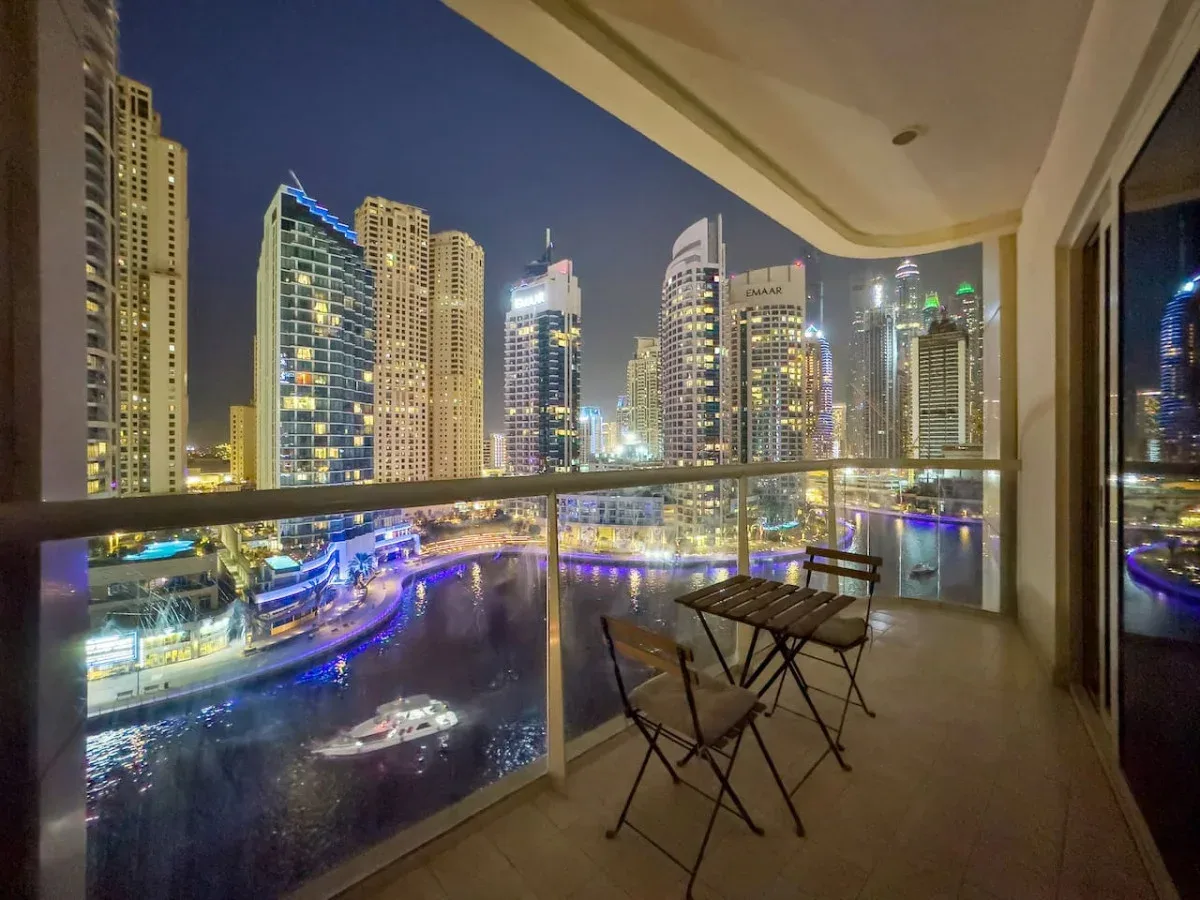 A balcony seating area overlooks a brightly lit waterfront cityscape at night.