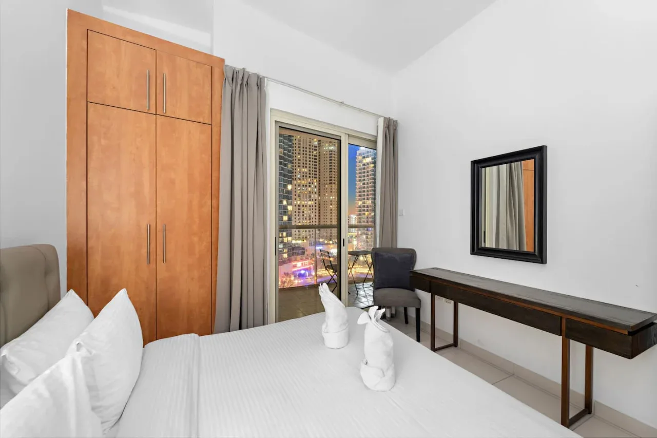 A bright bedroom with a bed, wooden wardrobe, and large window overlooking city lights.