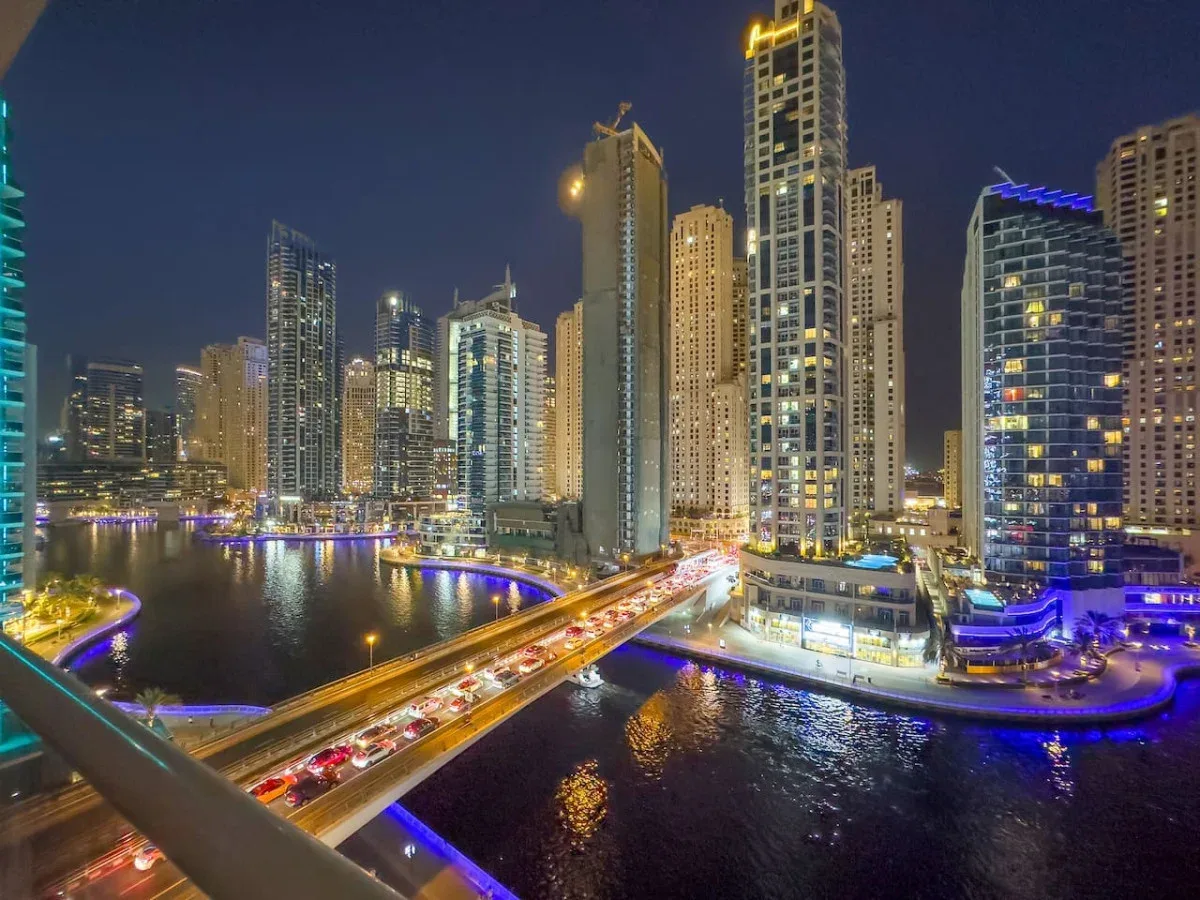A nighttime city skyline rises beside a waterway crossed by a brightly lit bridge.
