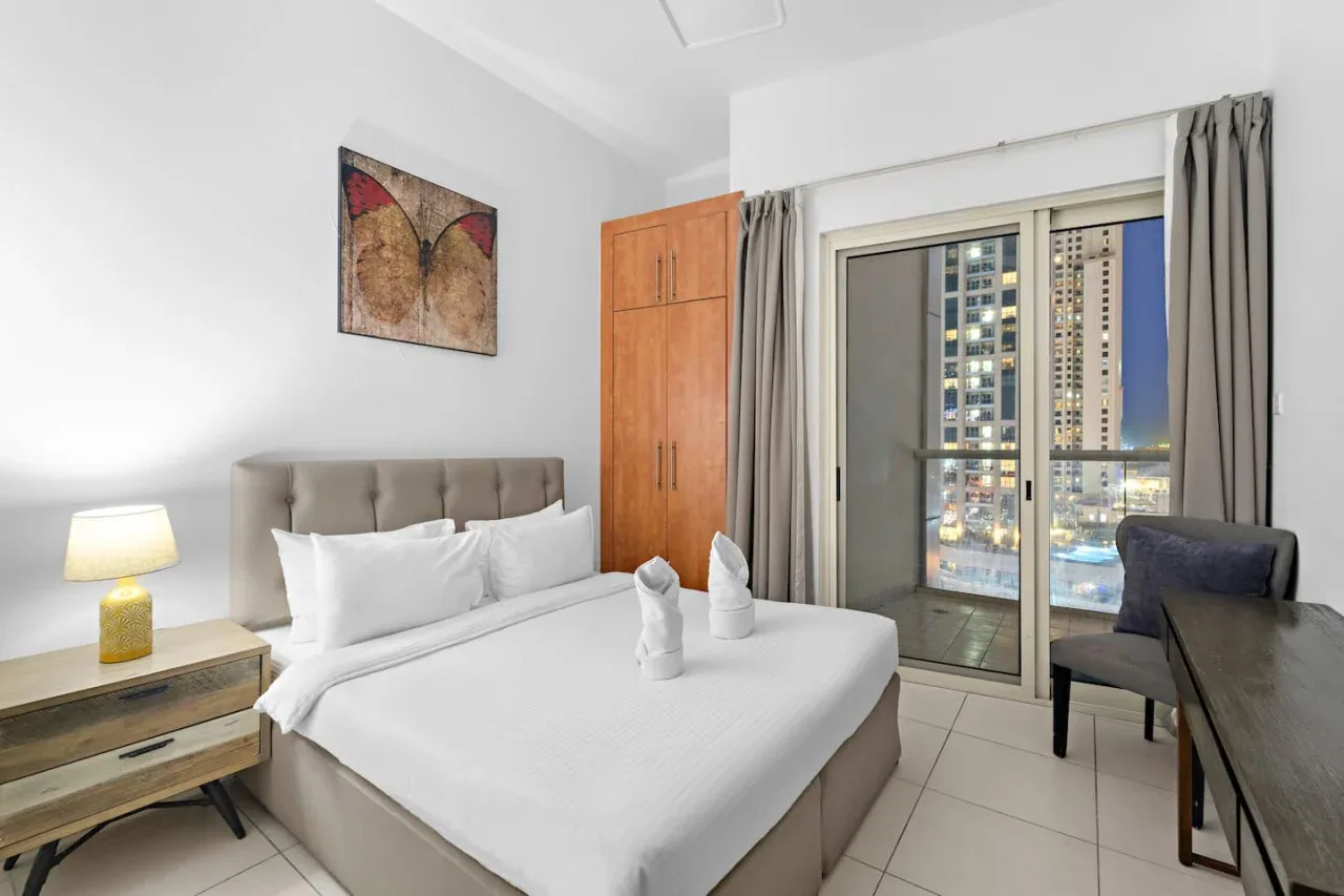A bedroom with a neatly made bed beside a window overlooking illuminated buildings.