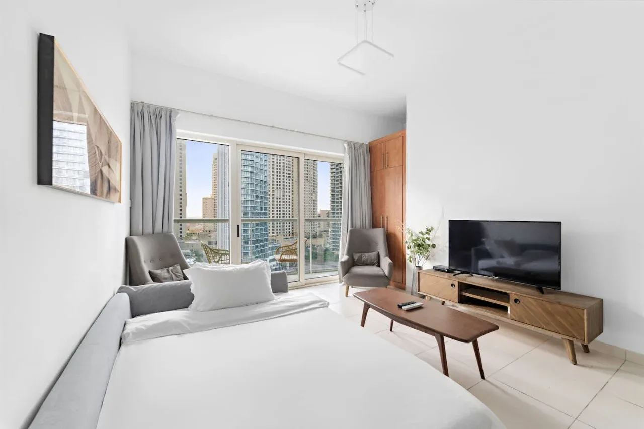 A bright living room with seating, a television, and large windows overlooking nearby buildings.