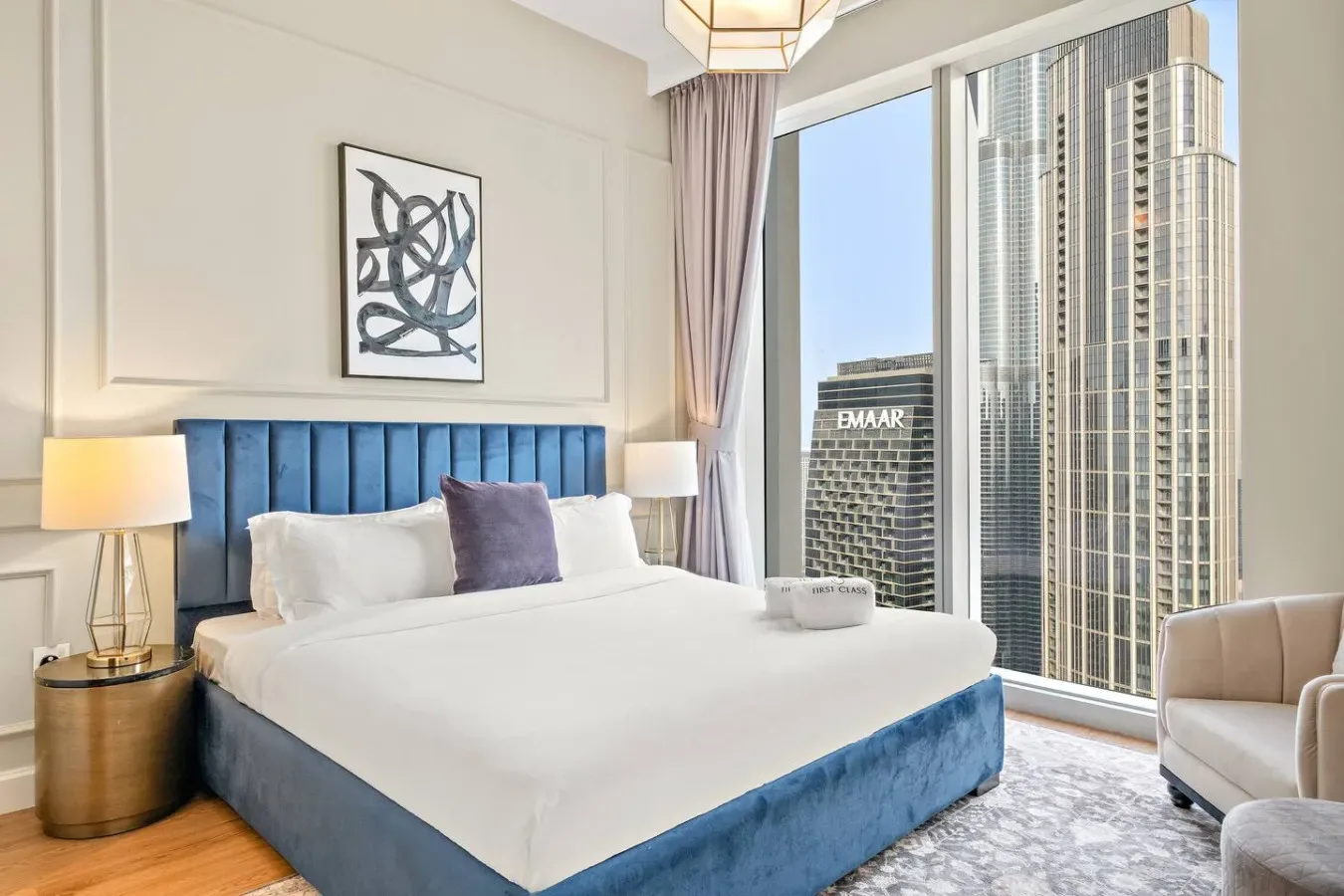 Un dormitorio con una cama tapizada en azul junto a grandes ventanales con vistas a edificios cercanos.