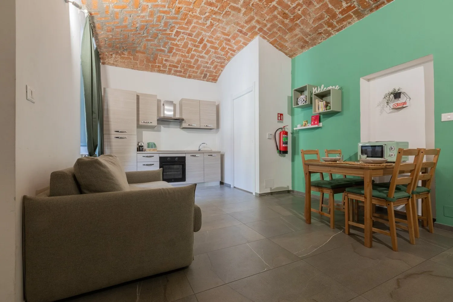 A living and dining area with a kitchen, wooden table, and exposed brick ceiling.