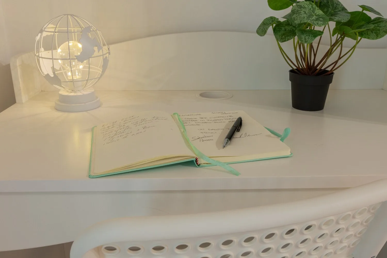 A desk holds an open notebook, a pen, a globe-shaped lamp, and a potted plant.