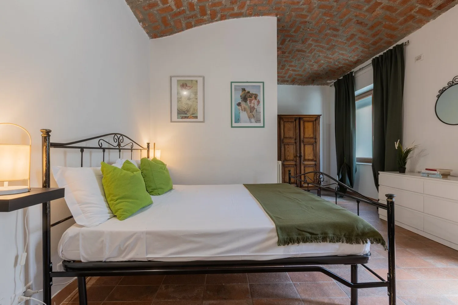 A bedroom with a metal bed, green pillows, and a brick vaulted ceiling.