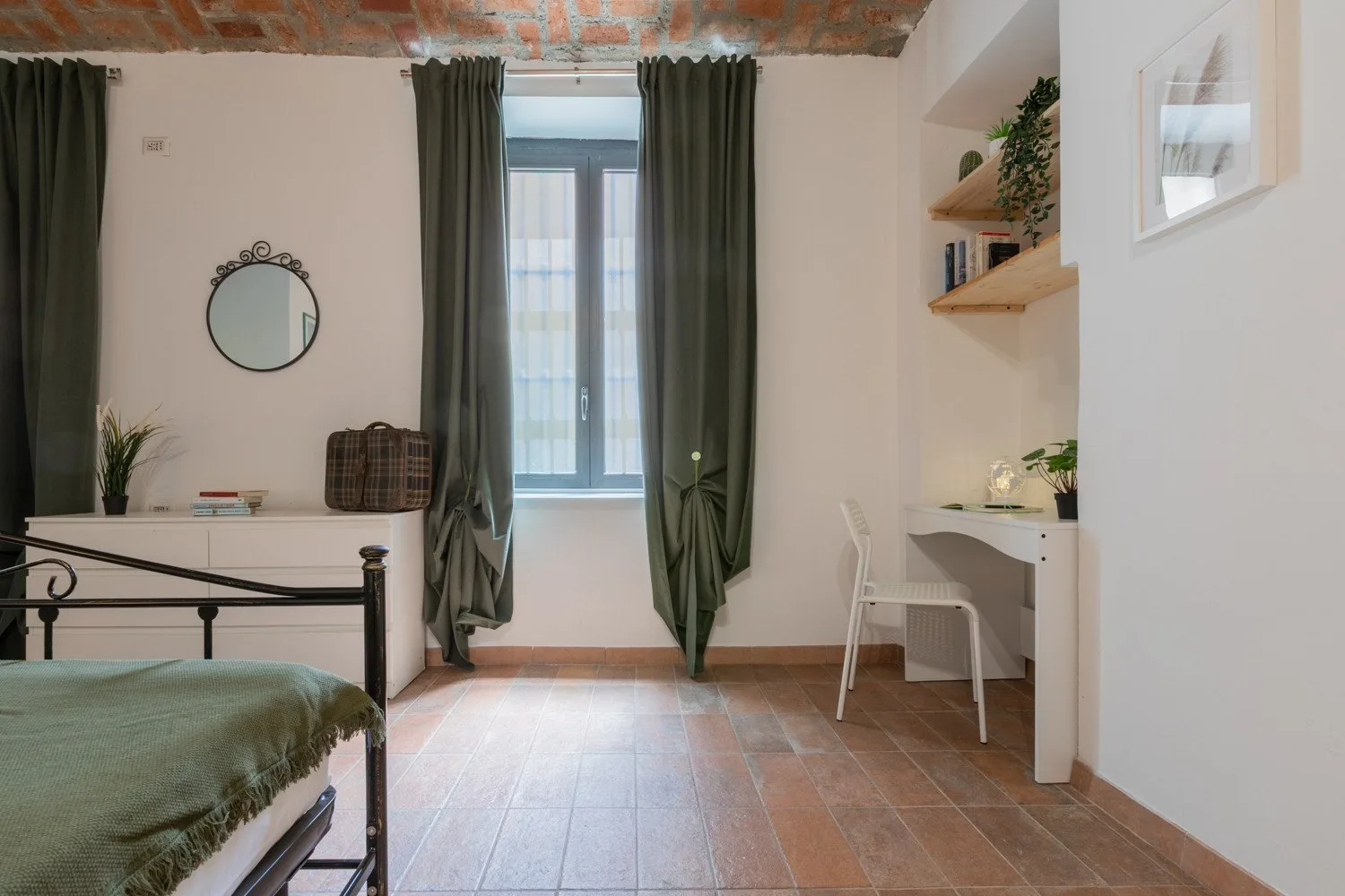 A bedroom with a bed, desk, and tall window framed by dark curtains.