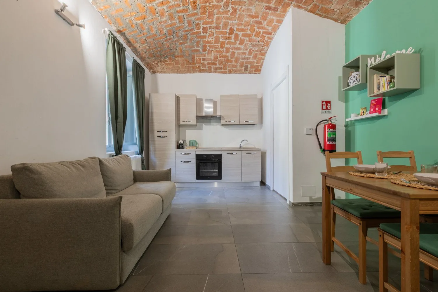 A living and dining area with a sofa, table, and compact kitchen beneath a brick ceiling.