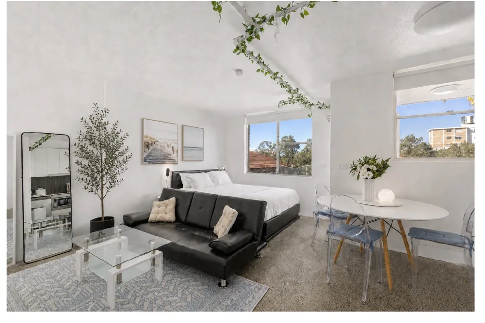 A bright studio room with a bed, dark seating, and a small dining table.