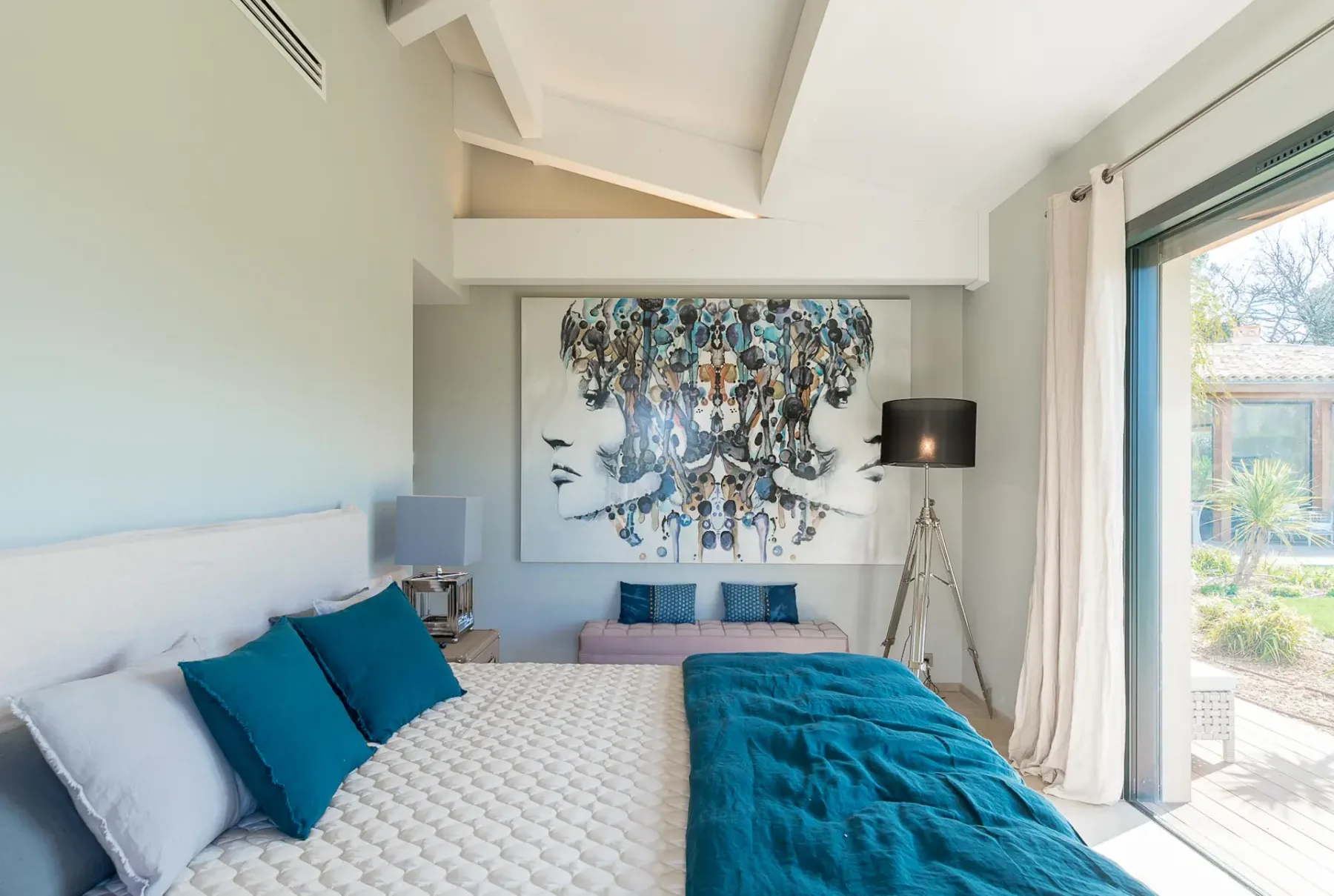 Cama doble con una obra de arte en la pared a la izquierda y una puerta corredera de cristal enfrente