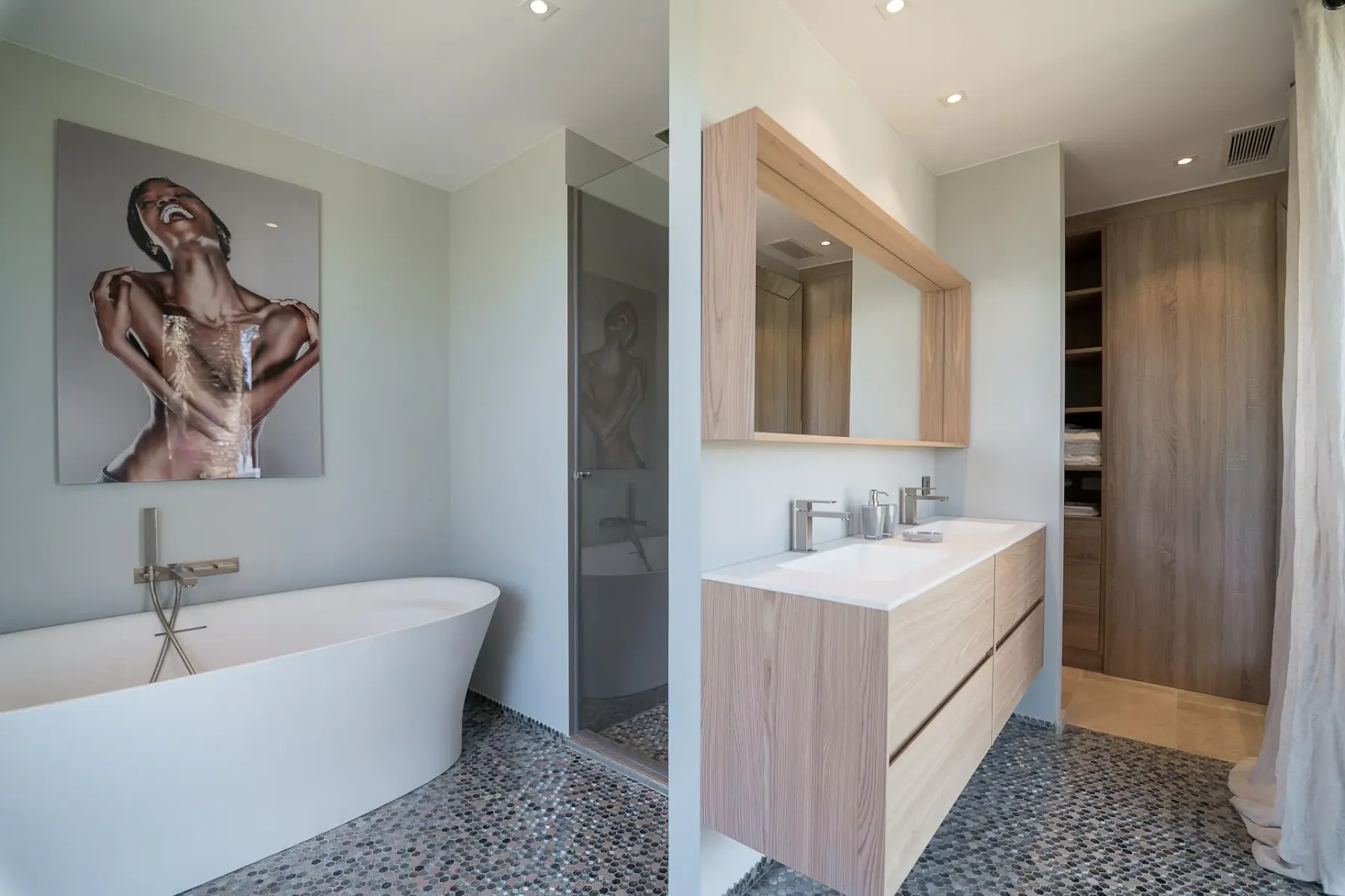 Baño moderno con lavabo y espejo, una pared divisoria con una bañera y cuadros colgados detrás.